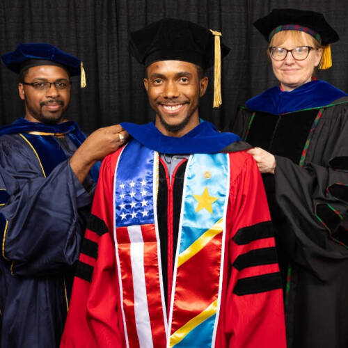 A smiling graduate in a red doctoral gown with stoles featuring the American and Congolese flags is being hooded by two faculty members in academic regalia, all standing in front of a black curtain backdrop.